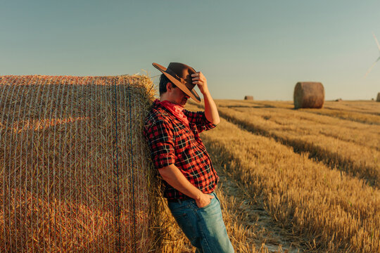 Stylish cowboy in flannel shirt and denim jeans with hat at sunset in wheat field