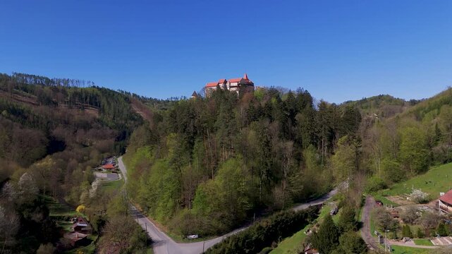 Drone ascends over the lush forest canopy, dramatically revealing the majestic Pernstein Castle perched on its rock above Nedvedice, South Moravia, Czech Republic, under a clear blue spring sky.