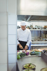 Male chef slicing vegetables on wooden cutting board in professional kitchen near stainless pots
