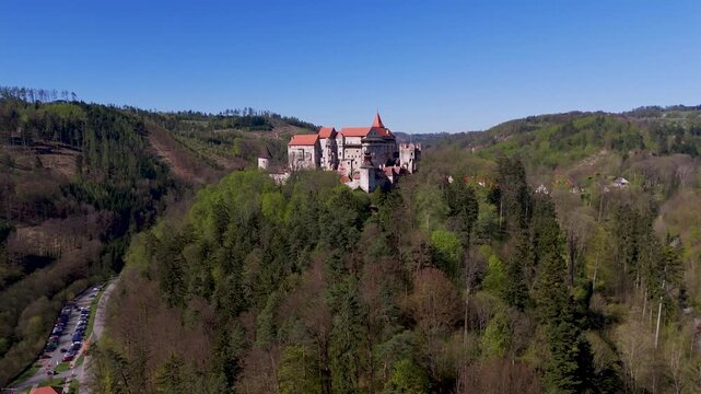 Aerial drone orbit reveals the majestic Pernstein Castle, a Gothic and Renaissance fortress perched dramatically on a massive rock formation overlooking Nedvedice, South Moravia, Czechia.