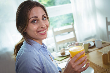 Woman in 30s sitting at dining table holding glass of orange juice near window, copy space