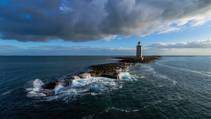 Aerial view of lighthouse on rocky outcrop surrounded by ocean waves under cloudy skies at daytime
