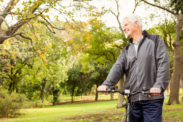 Senior man standing holding bicycle with front light and smiling looking right in park, copy space
