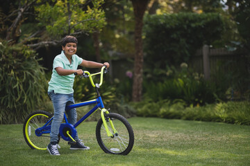 Obraz premium Asian boy smiling while straddling blue bicycle with neon yellow forks on garden lawn