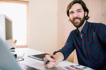 Bearded man in plaid shirt smiling toward camera while holding pen over printouts at home office