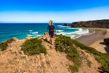 Beautiful mature woman tourist walking on cliff overlooking Odeceixe beach and ocean in summertime. Algarve Portugal. Back view