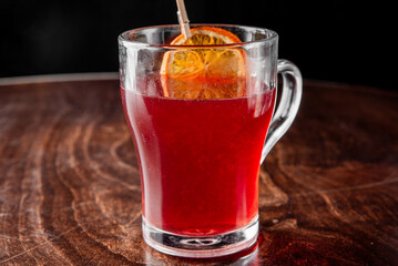 Glass mug with red herbal tea, garnished with dried orange slice and wooden stirrer, placed on rustic wooden surface. Cozy, aromatic drink perfect for seasonal or wellness themes.