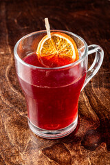 Glass mug with red herbal tea, garnished with dried orange slice and wooden stirrer, placed on rustic wooden surface. Cozy, aromatic drink perfect for seasonal or wellness themes.