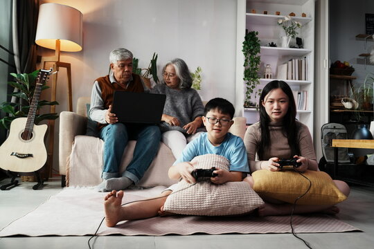 Asian teenager grandson and granddaughter sitting on floor holding joystick remote control game playing esports game having fun happy grandpa and grandma sitting on sofa in living room at home family