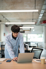Smiling glasses asian man standing leaning over while looking using laptop on wooden table in cafe.
