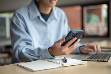 Asian man looking and holding smartphone over notebook aside laptop while sit at wood table in cafe