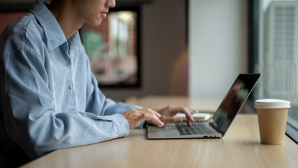 Asian man looking and typing on laptop aside book and coffee while sitting at wooden table in a cafe