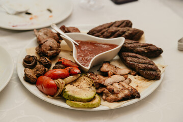 On a banquet table covered with a white tablecloth stands a white plate with meat, vegetables and sauce in a separate sauce pan