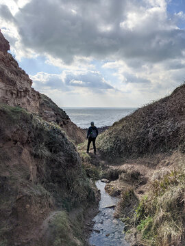 Adventurous Hiker on Cliffside Path Overlooking Ocean with Dramatic Sky, Rocky Terrain and Winding Stream in Sussex, England