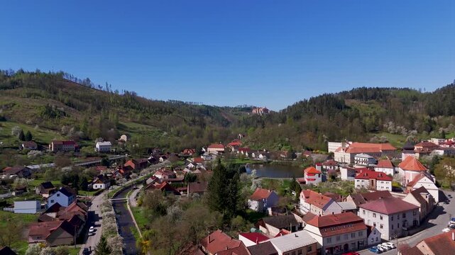 Aerial drone flight sweeping over the traditional rooftops of Nedvedice village, South Moravia, towards the majestic Pernstein Castle nestled among the rolling green spring hills of Czechia.