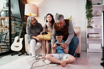 A happy grandparents family with their granddaughter and grandson in the living room of their home. Grandfather holds the joystick and grandson plays games together.