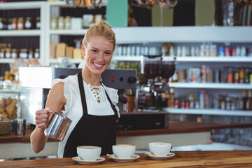Female barista wearing apron holding pitcher pouring milk into espresso cups behind counter at cafe