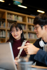 Glasses asian woman student looking at laptop of a man friend while sitting at table in library.