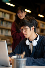 Glasses asian man student looking at laptop with a woman friend standing beside table in library.