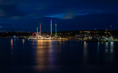 Obraz premium A panoramic night view of Djurgården, Stockholm. Attractions like Gröna Lund, Skansen, Kaknästornet, and the Vasa Museum illuminate the night and reflect in the water.