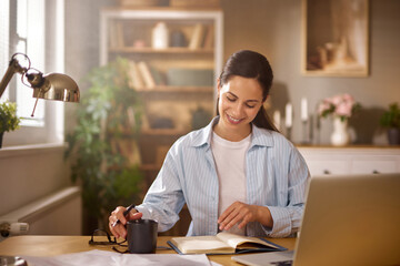 Fototapeta premium A woman is sitting at her home office desk, writing in a notebook and sipping coffee from a mug, with a laptop open in front of her and a warm, cozy atmosphere surrounding her.