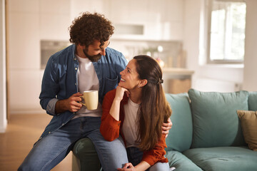 A man and woman share a warm moment in a stylish living room. They smile at each other, holding a coffee mug, surrounded by soft furnishings and natural light.