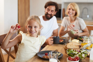 A cheerful girl holds a cherry tomato while her parents smile and share a meal. The bright kitchen setting enhances the warm atmosphere of family bonding during breakfast.