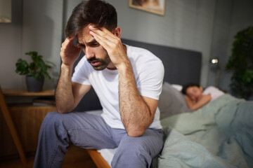 A man sits on the edge of the bed with his head held in his hands, distressed by relationship troubles. His partner lies asleep beside him, unaware of his turmoil.