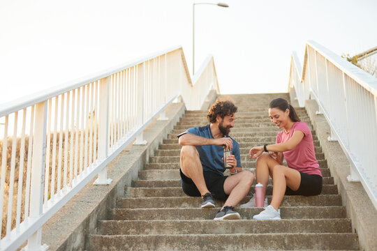 Two individuals are sitting on outdoor steps, enjoying a moment of relaxation after exercising. They are smiling and discussing their fitness trackers under the warm glow of sunset.