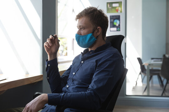 Man in 30s in office chair wearing blue face mask, holding pen and gazing out window