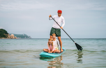 Father paddleboarding with two kids in tropical waters. Smiling, laughing and splashing joy of active bonding and beach adventure on summer family vacation.