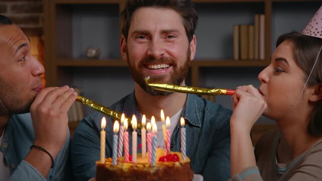 Happy birthday adult Caucasian man with cake burning candles smiling looking at camera two multiethnic guy woman blowing in party blowers whistles at home three multiracial friends celebrate together