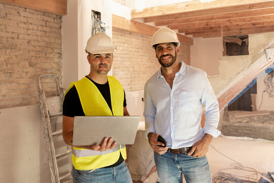Two male engineers and architects with laptop standing at building site smiling at camera