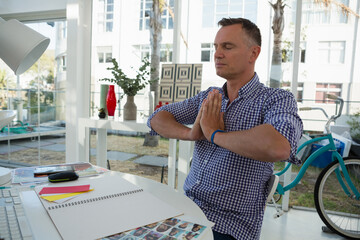 Meditating man holding prayer pose at home office desk, with sketchbook and bicycle, copy space