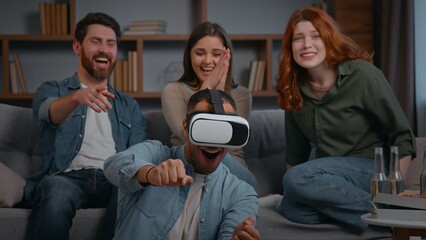 Four multiracial friends at home playing VR game laughing at home party three buddies supporting man in virtual reality helmet play smiling together gaming having fun interactive women men on couch