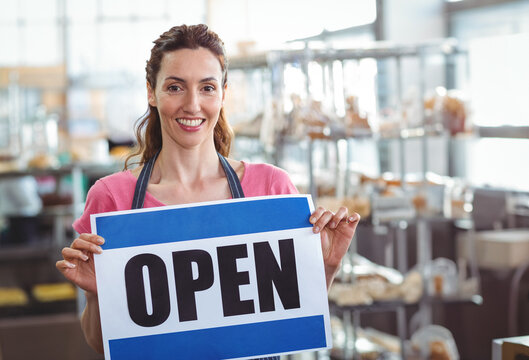 Smiling woman baker holding OPEN sign inside bakery, with metal racks and pastries - Powered by Adobe