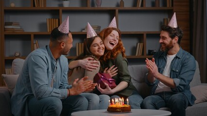 Four multiracial friends celebrating birthday party presenting present Caucasian woman receive gift from diverse friendly buddies men women cuddling applauding happy smiling congratulate event at home