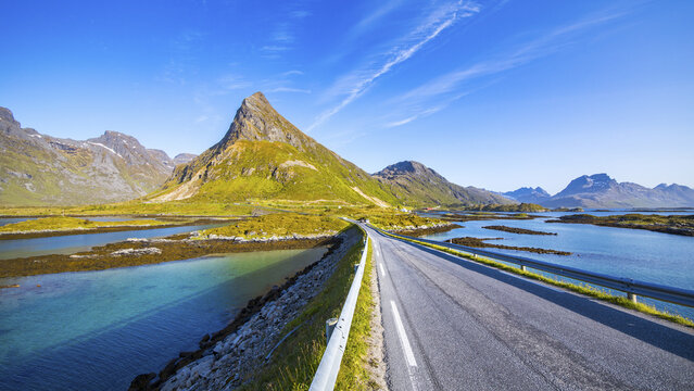 View of a long asphalt road cuts through the serene turquoise waters, leading towards a towering, green-capped mountain under a clear blue sky, Fredvang, Lofoten, Norway.