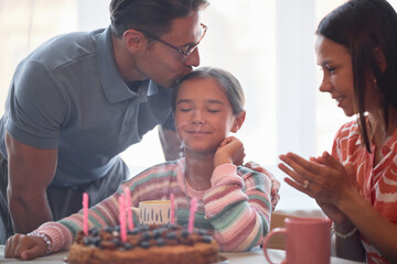 Caucasian man kissing head of smiling child girl with closed eyes while sitting at table with birthday cake and candles, young adult woman clapping hands and watching celebration