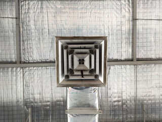 A detailed view of an air conditioning vent mounted on a warehouse ceiling. The metallic vent has a tiered, square design, while the ceiling is covered in reflective, crumpled foil insulation.