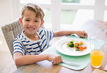 Child eating steamed vegetables with fork and knife and drinking orange juice at home dining table