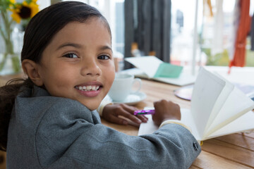 Writing girl child using purple pen over open notebook at table near window, with sunflower vase