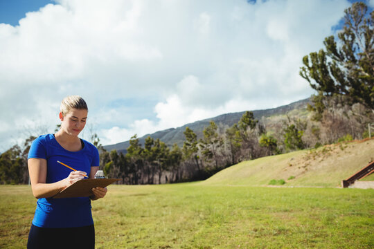Woman writing on clipboard in grassy rural field near rolling hills under partly cloudy sky - Powered by Adobe