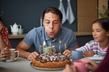 Caucasian young adult man blowing out candles on birthday cake while sitting at table with...