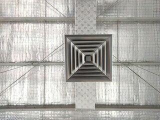 A detailed view of an air conditioning vent mounted on a warehouse ceiling. The metallic vent has a...