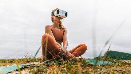 Low angle view of woman wearing VR headset sitting cross-legged on yoga mat in nature. Symbol of harmony between technology and natural environment.