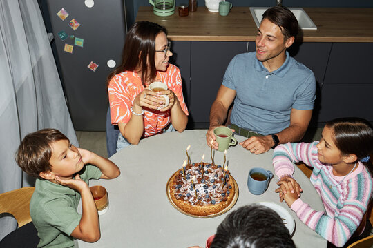 Caucasian young adult man and woman sitting at round table with two Caucasian children, celebrating birthday with cake and candles, holding mugs, engaging in conversation