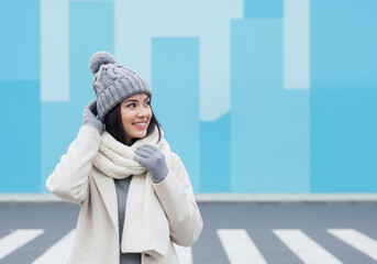 Happy young woman in stylish winter clothes. Smiling person wearing a warm beanie and scarf outdoors in the city. Cold weather fashion with blue copy space