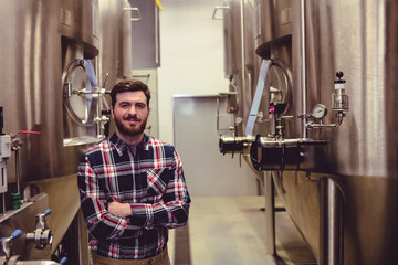 Standing male in his twenties crossing arms at industrial brewery, with fermentation tanks
