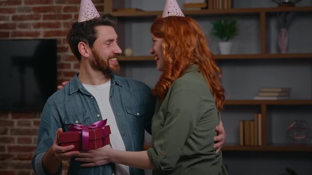 Two Caucasian friends couple at home party birthday congratulation woman congratulate man giving present box festive gift surprise excited male smiling cuddling love female celebrating together family